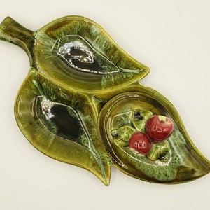 May include: A green ceramic serving dish shaped like a leaf with three separate compartments. The dish has a glossy glaze with brown and silver accents. The center compartment features a raised design of red cherries.