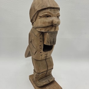May include: A hand-carved wooden nutcracker in the shape of a man with a hat and beard. The nutcracker is standing on a rectangular wooden base. The wood has a natural, light brown color.