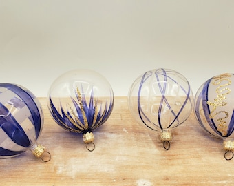 Pode incluir: Quatro enfeites de Natal de vidro com desenhos pintados em azul e dourado. Os enfeites são de vidro transparente com listras azuis, redemoinhos azuis e dourados, estrelas azuis e douradas e listras azuis com letras douradas.