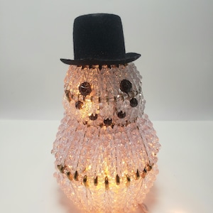 May include: A white, beaded snowman with a black top hat and a string of lights. The snowman is lit up from the inside, creating a warm glow.