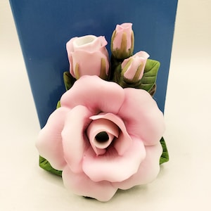 May include: A ceramic rose bud vase with a pink rose and two smaller pink rose buds. The vase is decorated with green leaves.
