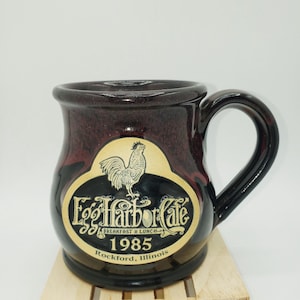 May include: A black and red ceramic mug with a gold rooster and the text "Egg Harbor Cafe Breakfast & Lunch - 1985 Rockford, Illinois" on the front.