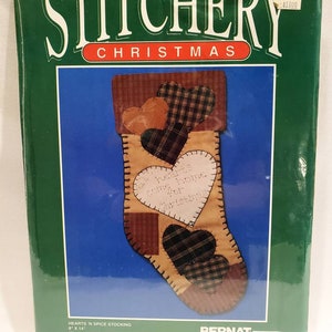 May include: A Bernat Stitchery Christmas stocking kit featuring a heart-shaped design with the text "Our hearts come home for Christmas". The kit includes natural muslin, fabric prints, batting, fusible webbing, needles, full-size patterns, directions, and floss and pearl cotton thread. The kit is 8 inches by 14 inches.