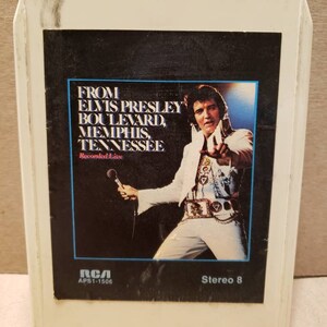 May include: A white cassette tape with a black label featuring a photo of Elvis Presley performing on stage. The label reads "From Elvis Presley Boulevard, Memphis, Tennessee" and "Recorded Live". The cassette tape is labeled "RCA APS1-1506" and "Stereo 8".