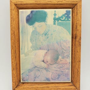 May include: A framed print featuring a mother and child. The artwork depicts a woman in a light-colored dress and a baby lying on a patterned blanket. The frame is made of light brown wood.