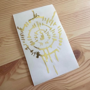 以下が含まれることがあります： 白い背景に抽象的なデザインが施された、金色のステッカー。 デザインは、放射線状の線と様式化された形状を持つ中心の円形要素を特徴としています。 ステッカーは長方形で、光沢のある仕上がりです。