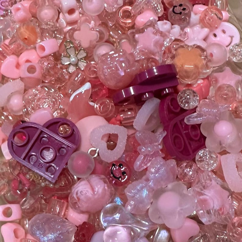 May include: A collection of pink and clear beads and charms. The assortment includes heart shapes, stars, flowers, and other decorative elements. Some pieces have a glossy finish, while others are translucent.