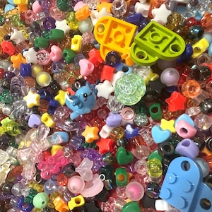 May include: A vibrant collection of craft beads and small plastic shapes. The assortment features stars, hearts, and other decorative elements in various colours, including red, yellow, blue, and green. Some pieces are transparent.