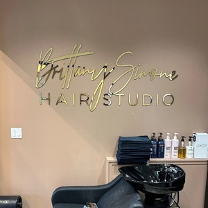 May include: A gold sign with the text "Brittany Simone Hair Studio" on a light pink wall. The sign is above a black salon chair and a black sink.