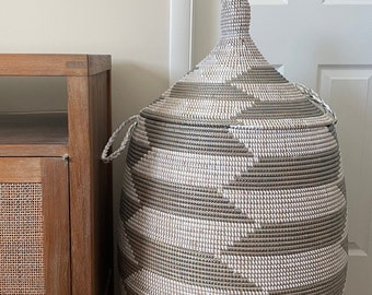 May include: A large, woven storage basket with a lid. The basket is mainly white and grey, with a geometric pattern. It has a rounded shape and a handle on the lid. The basket is next to a wooden cabinet.