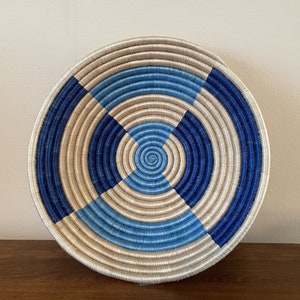 May include: A round woven basket with a concentric design. The basket features alternating sections of light blue, dark blue, and natural beige tones. The basket is circular and appears to be made of tightly woven fibers.