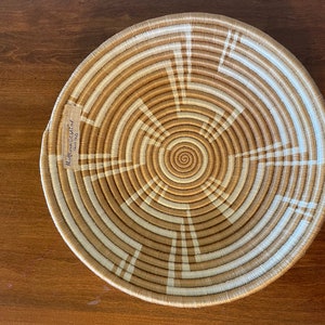 May include: A large, round woven basket with a tan and white pattern. The basket has a spiral design in the center and is made of natural materials.