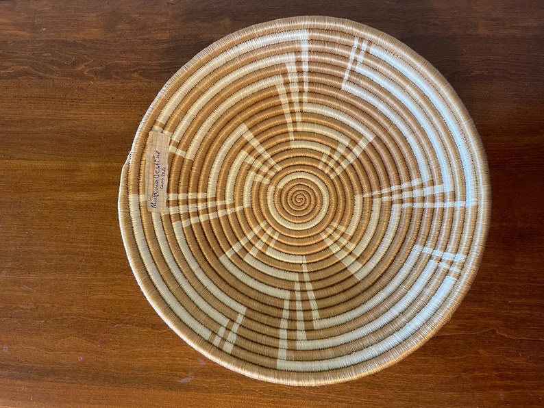 May include: A large, round woven basket with a tan and white pattern. The basket has a spiral design in the center and is made of natural materials.