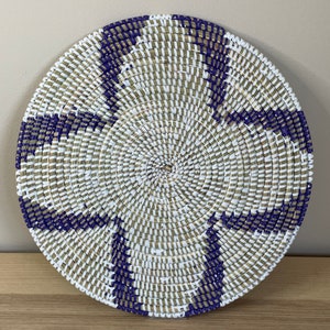 Pode incluir: Um pendurado de parede redondo tecido com um padrão geométrico branco e roxo. O design tecido é feito de fibras naturais e tem uma forma circular.