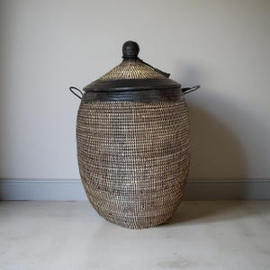 Op de afbeelding: Een grote, geweven mand met een deksel. De mand heeft een cilindrische vorm met een afgeronde bodem en twee handvatten. Het deksel is conisch en heeft een kleine, ronde knop. De mand is gemaakt van natuurlijke vezels in beige en zwart.
