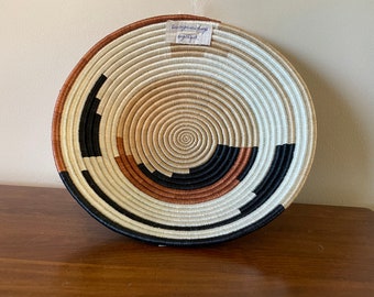 Puede incluir: Una cesta grande y redonda tejida con un patrón circular de rayas negras, blancas y marrones. La cesta está hecha de fibras naturales y tiene un aspecto rústico y hecho a mano.
