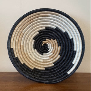 May include: A large, round woven basket with a black and white spiral pattern. The basket is made of natural fibers and has a rustic, handmade look.