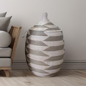 Può includere: Un grande cesto portaoggetti intrecciato con coperchio. Il cesto è principalmente bianco e grigio, con un motivo geometrico. Ha due manici e una forma arrotondata. Il cesto è accanto a una sedia di legno e a un cuscino grigio.