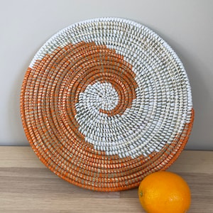 Könnte beinhalten: Ein runder gewebter Wandbehang mit einem Spiralmuster in Orange und Weiß. Der Wandbehang ist aus natürlichen Materialien gefertigt und hat einen rustikalen, bohemian Stil.