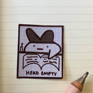 May include: A square sticker with a cartoon character wearing a hat, reading a book. The words "HEAD EMPTY" are printed below the book. The sticker is outlined in brown and set against a grid background.
