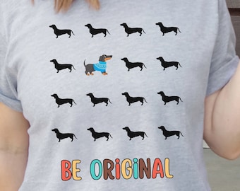 May include: A grey t-shirt with a black dachshund wearing a blue jumper in the centre of a grid of black dachshunds. The text "BE ORIGINAL" is printed below the grid in rainbow colours.