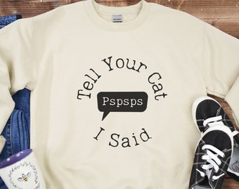 Può includere: Una felpa color crema con una grafica circolare nera che dice "Tell Your Cat I Said" con un fumetto che dice "Pspsps".