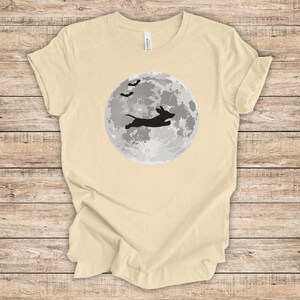 May include: A light beige t-shirt with a black silhouette of a dachshund jumping in front of a full moon with bats flying above.