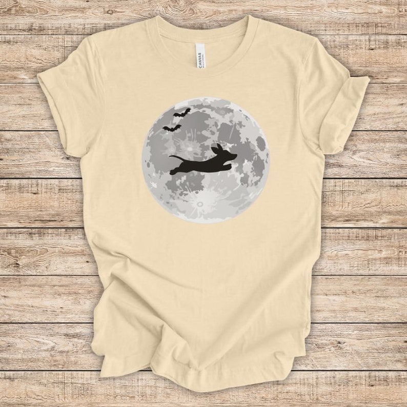 May include: A light beige t-shirt with a black silhouette of a dachshund jumping in front of a full moon with bats flying above.