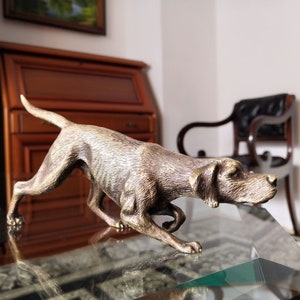 May include: A bronze-coloured dog sculpture in a running pose. The dog has a long snout, floppy ears, and a slender body. The sculpture is on a glass surface, with a blurred background of wooden furniture and a chair.