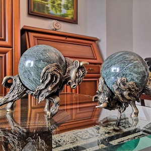 Könnte beinhalten: Zwei bronzene Stier-Skulpturen, die grüne Marmorkugeln halten. Die Stiere stehen auf einem Glastisch mit einer Holzoberfläche darunter.