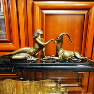 Könnte beinhalten: Eine Bronzeskulptur einer Frau und einer Gazelle auf einem schwarzen Marmorsockel. Die Frau liegt mit ausgestrecktem Arm in Richtung der Gazelle, die mit dem Kopf zur Frau gewandt daliegt.
