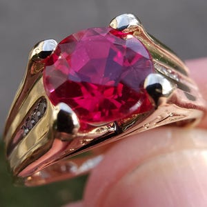 May include: A gold-toned ring featuring a large, faceted, deep red gemstone as the centerpiece. The ring has a classic design with a band that widens towards the top, and small accent stones are set on the sides. The gemstone is the focal point.