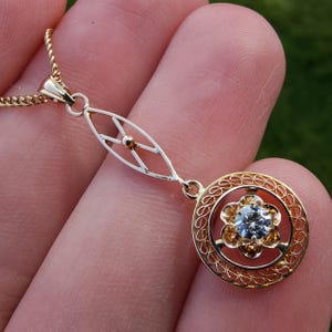 Peut inclure: Un collier pendentif en or doté d'un cadre circulaire en filigrane avec un sertissage central en forme de fleur contenant une pierre précieuse ronde et claire. Le pendentif est relié à une fine chaîne en or par un connecteur marquise ajouré.