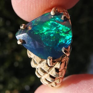 Op de afbeelding: Een gouden ring met een grote, peervormige opaal. De opaal vertoont een levendig kleurenspel, met flitsen van groen en blauw tegen een donkere achtergrond. De band van de ring heeft een textuur, geweven ontwerp.