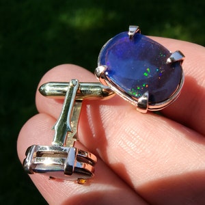 May include: A pair of gold cufflinks with a blue opal gemstone set in each. The cufflinks have a simple design with a smooth, polished finish.