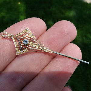 May include: A gold pendant with a diamond-like center stone, small pearls, and leaf accents. The pendant has an open filigree design and a long, thin bar extending from the bottom. The pendant is held in a hand against a blurred green background.