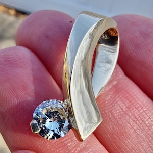 Può includere: Un anello d'oro con una singola pietra preziosa grande e trasparente. L'anello è progettato con una fascia curva che si avvolge attorno al dito.