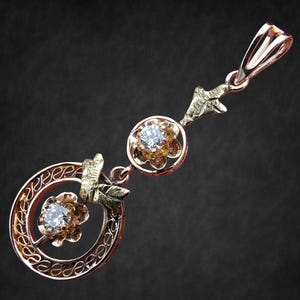 May include: A rose gold pendant with two flower-shaped settings, each holding a clear gemstone. The pendant features intricate filigree and leaf accents, with a loop at the top for a chain. The design is set against a dark background.