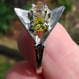 Op de afbeelding: Een zilveren ring met een grote, lichtgele edelsteen boven een kleinere, heldergele steen. De ring heeft een uniek, vleugelachtig ontwerp aan weerszijden van de edelstenen. De achtergrond is wazig en groen.