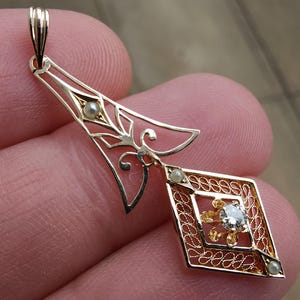 May include: A gold pendant with an ornate filigree design. The pendant features a diamond-shaped element with a central gemstone and small pearls. The top section has a decorative openwork design. The pendant is held up by a small loop.