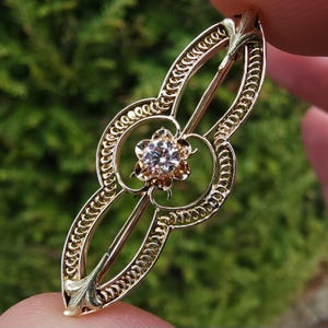 Può includere: Un fermaglio in filigrana d'oro con un solo diamante rotondo al centro. Il fermaglio ha la forma di un fiore con dettagli intricati.