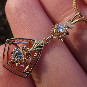 May include: A gold pendant necklace featuring two floral designs, each with a central, clear gemstone. The pendant has a geometric frame with a decorative leaf element. The necklace chain is a fine, gold link.