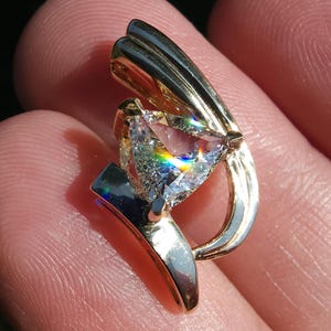 Könnte beinhalten: Ein goldener Anhänger mit einem großen, dreieckigen, klaren Edelstein. Der Edelstein reflektiert ein Regenbogenspektrum. Der Anhänger hat ein geschwungenes Design mit poliertem Finish und wird von goldenen Zinken gehalten.