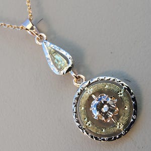 May include: A gold-toned pendant necklace with a teardrop-shaped bail and a circular pendant. The pendant features a central, clear gemstone surrounded by a gold filigree design and a decorative border. The chain is also gold-toned.