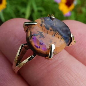 May include: A gold ring featuring a large, oval-shaped gemstone with a mix of colors, including brown, orange, and purple. The gemstone is held in place by four gold prongs. The ring's band is also gold.