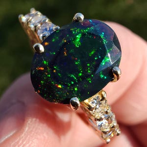 Op de afbeelding: Een gouden ring met een grote, ovaal geslepen zwarte opaal. De opaal vertoont groene, rode en gele flitsen. De gouden band is bezet met kleine, heldere edelstenen. Een opvallend sieraad voor speciale gelegenheden.