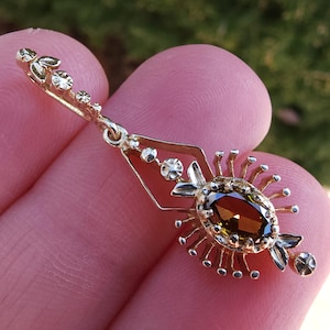 May include: A gold pendant with an oval, brown gemstone at its centre. The pendant has a detailed design with floral and geometric elements, and small, clear accent stones. The overall design is ornate and vintage-inspired.