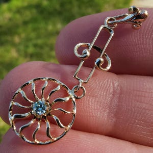 May include: Gold tone dangle earring with a blue gemstone in the center of a sunburst design. The earring has a music note shaped connector.