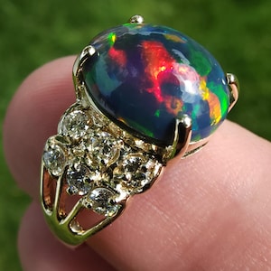 Op de afbeelding: Een gouden ring met een grote, ovale zwarte opaal die rode, groene en blauwe flitsen vertoont. De ring is versierd met kleine, heldere diamanten die in een cluster op de band zijn geplaatst. De opaal meet ongeveer 2 cm.