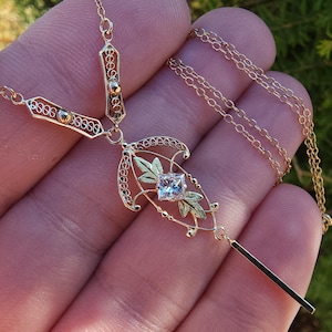 以下が含まれることがあります： 緑の葉で囲まれた正方形の透明な石を特徴とする繊細なペンダントが付いた金のネックレス。ペンダントは長く細い金のチェーンに取り付けられています。